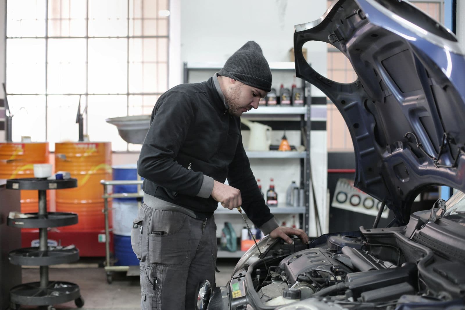 A mechanic in a black beanie and dark work clothes leaning under the open hood of a car inside a repair shop, with shelves of fluids behind him