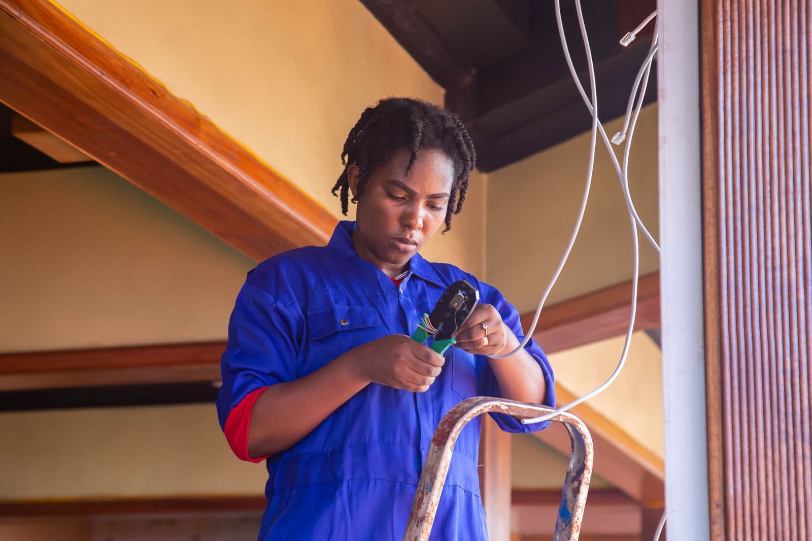 A technician in blue coveralls stands at a stepladder under a wooden ceiling beam, crimping a connector onto a white cable