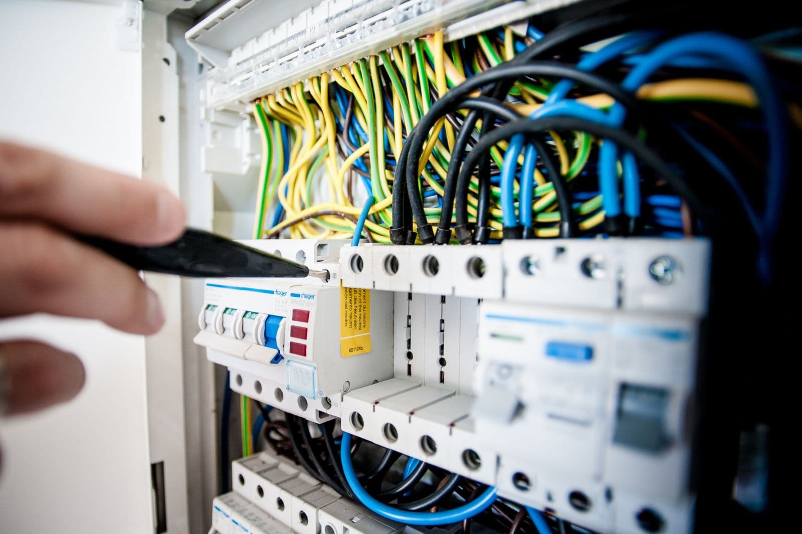 A hand holding a black tester against a row of circuit breakers in an open panel, with blue, black and yellow wiring behind