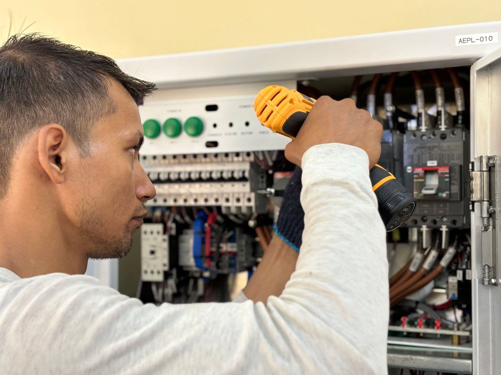 An electrician in a gray long-sleeve shirt using a yellow cordless drill inside an open electrical panel