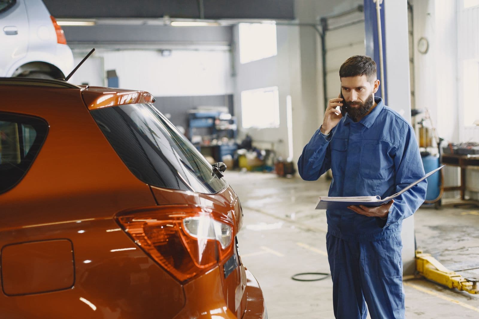 A bearded mechanic in blue coveralls on the phone, holding an open binder, standing behind an orange hatchback inside a repair shop with a lift and tool carts behind him