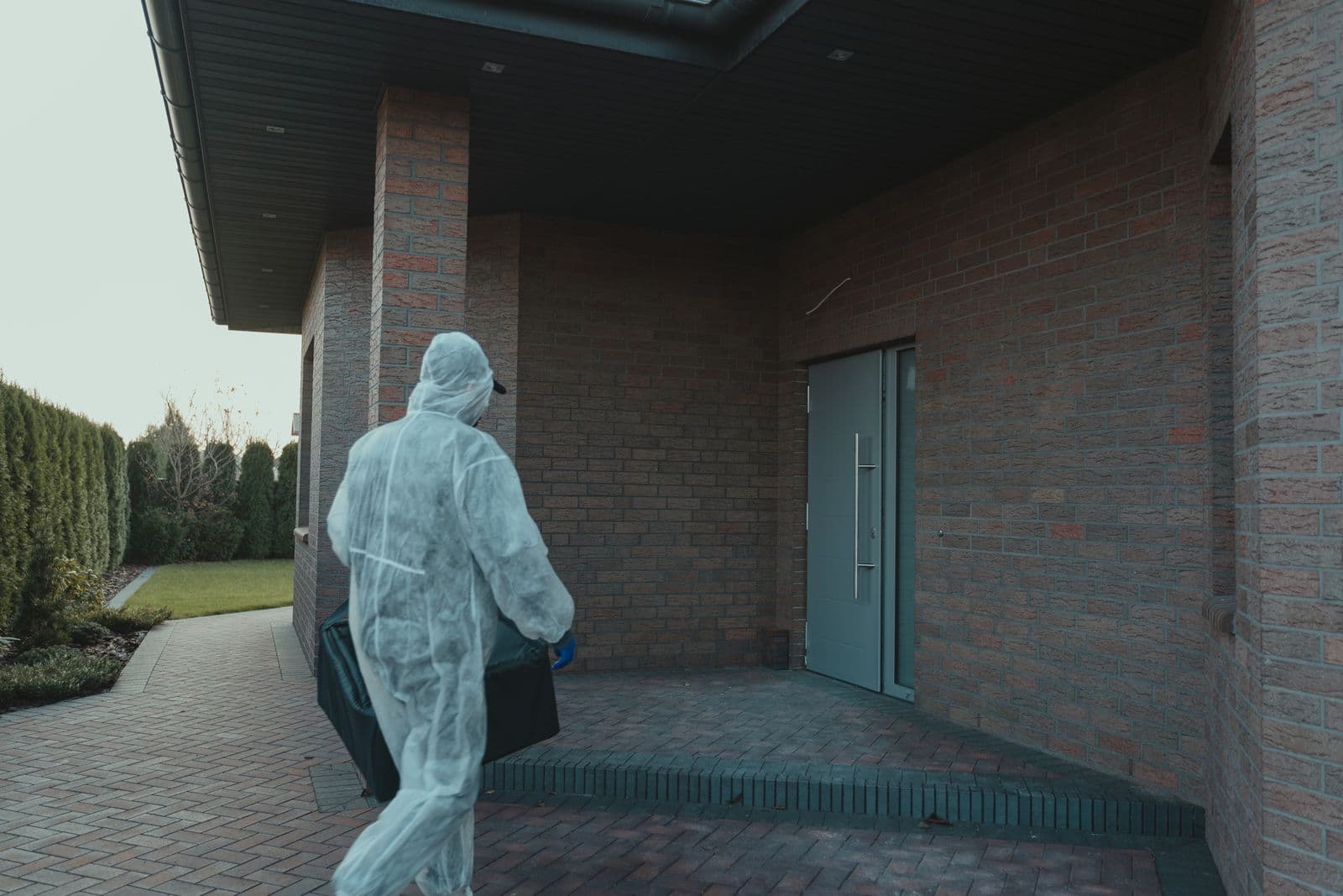 A person in a white protective coverall carries a black equipment case across a brick patio toward the front door of a brick house