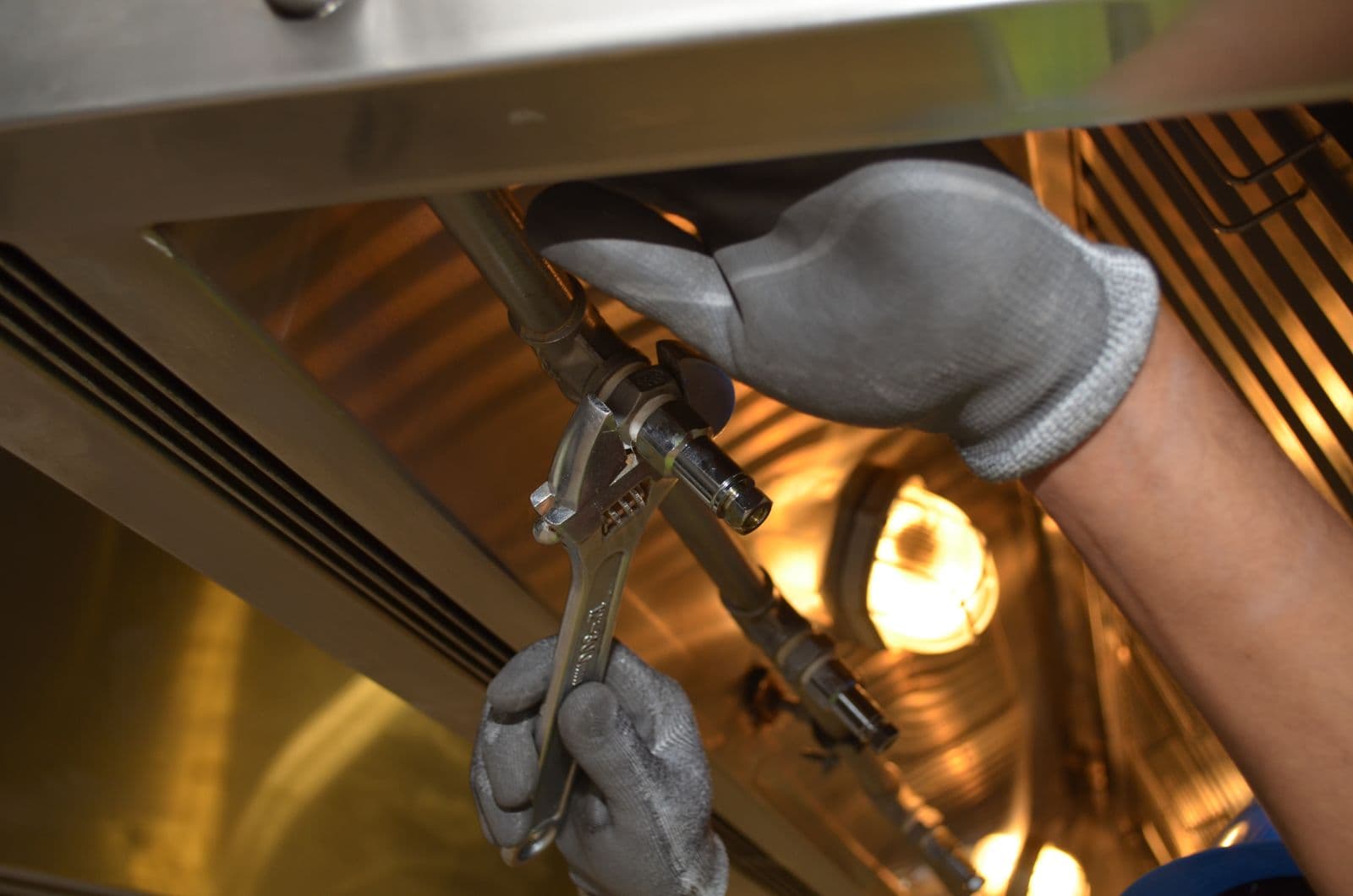 Two gloved hands tightening a pipe fitting with an adjustable wrench under a stainless steel hood