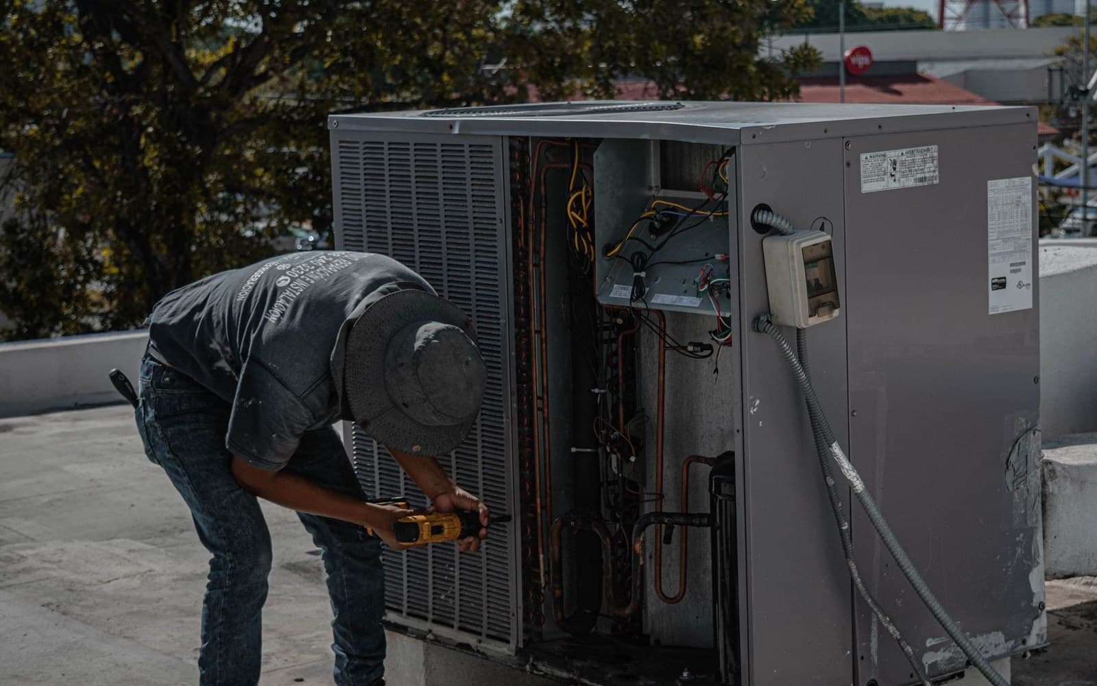 A technician in a bucket hat bends over an open rooftop air conditioning unit, holding a yellow cordless drill
