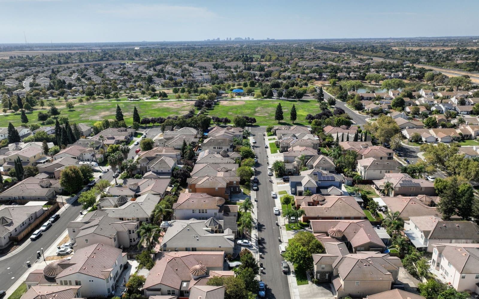 Aerial view of a suburban neighborhood of two-story tile-roof houses along a straight street, with a park and a distant city skyline