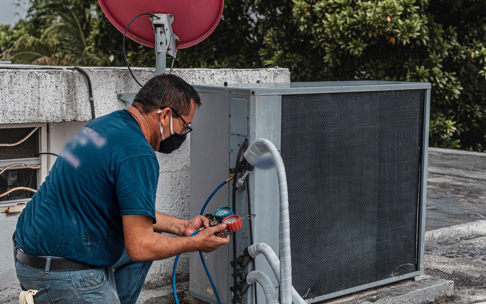 A technician in a blue T-shirt and face mask reading a manifold gauge hooked to an outdoor condenser on a flat roof, with trees behind him