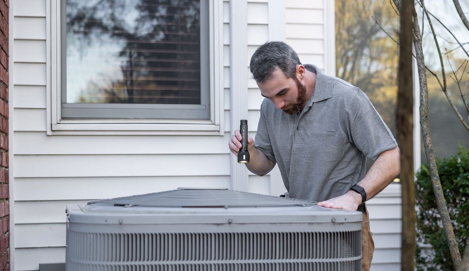 A technician with a flashlight checking the outdoor air conditioning unit beside a house with white siding