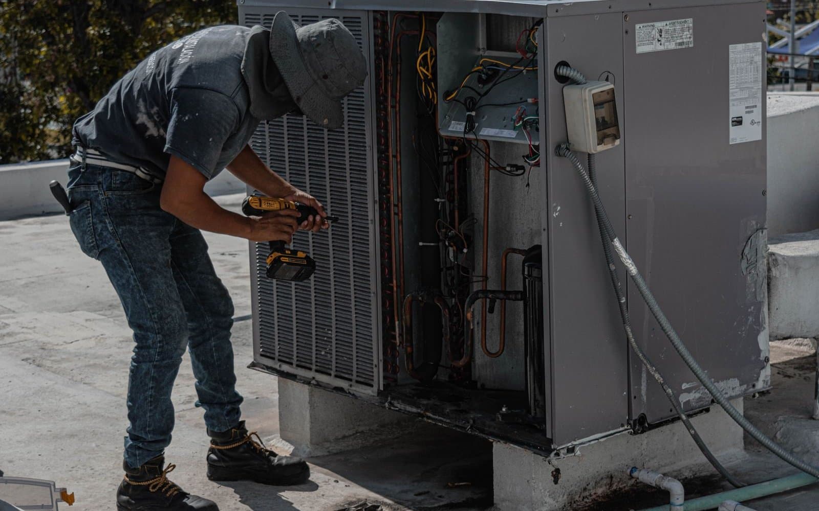 A technician in a bucket hat and gray T-shirt using a cordless drill on an open outdoor air conditioning unit on a flat rooftop