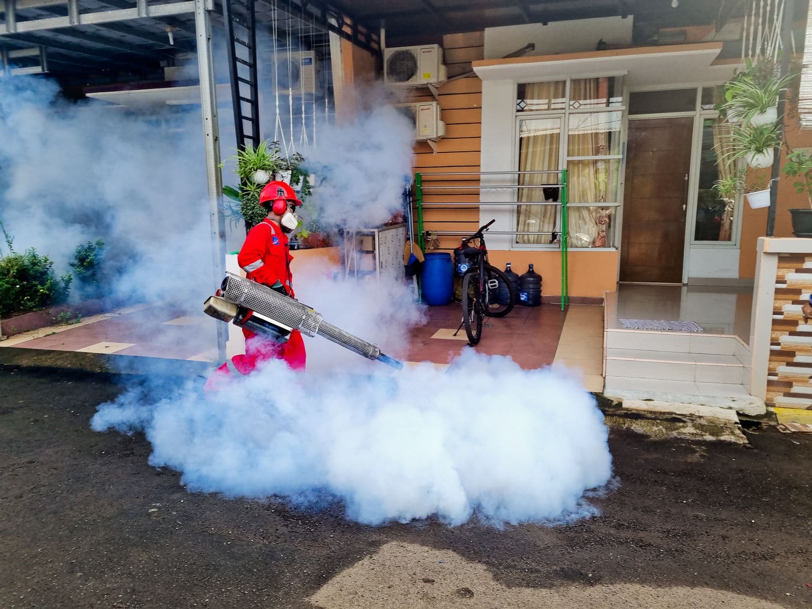 A pest control technician in a red suit, helmet and respirator fogging the front of a house with a thermal fogger