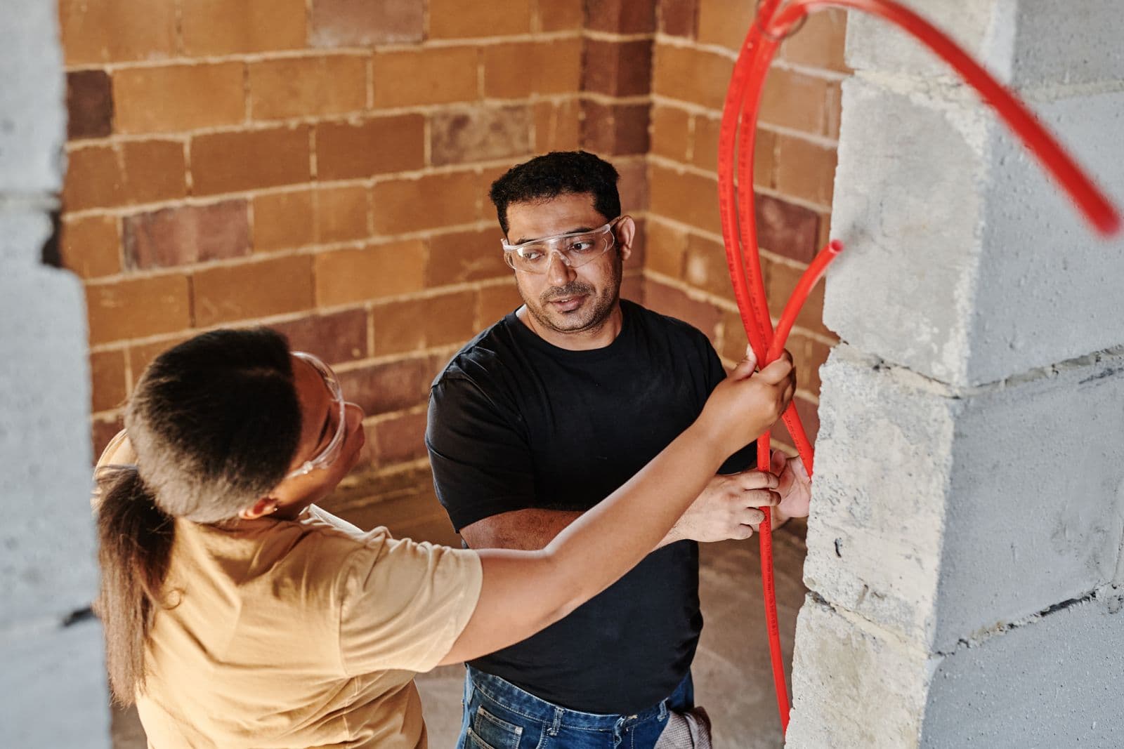 Two workers in safety glasses feeding red plumbing tubing through an unfinished block wall