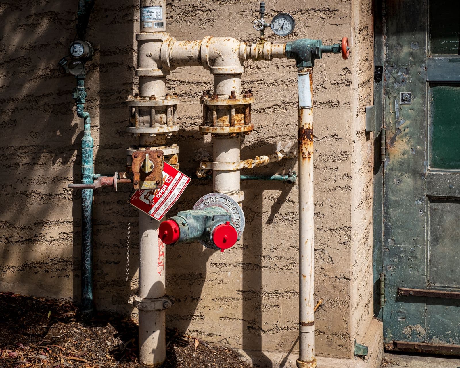Painted water and fire department connection pipes with a pressure gauge, a red valve wheel and a red tag on the outside wall of a stucco building